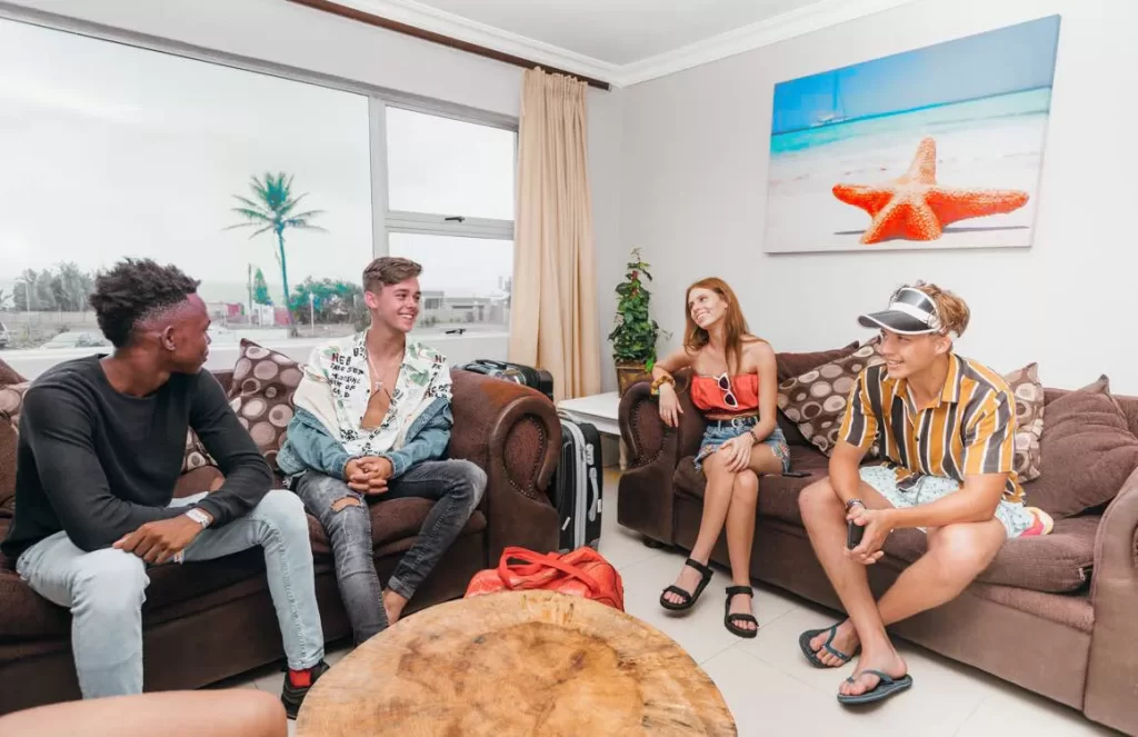 Four friends relaxing in a cozy lounge at Ballito Backpackers, with large windows showing a palm tree outside.